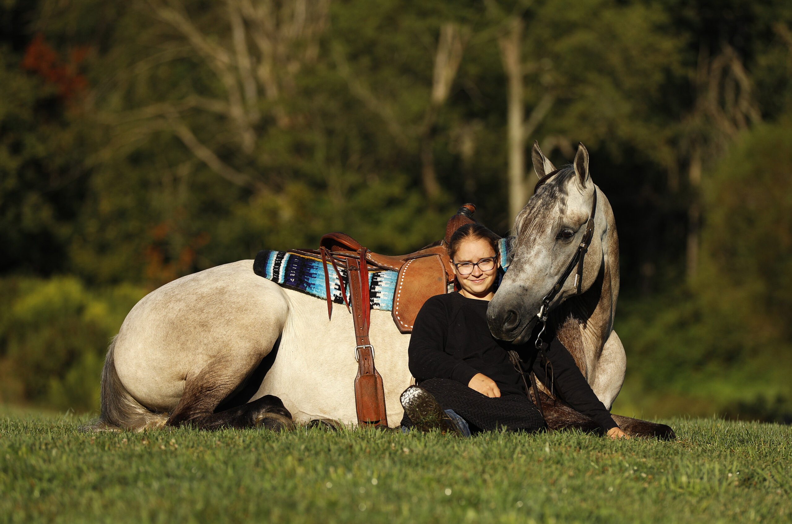 A woman with glasses sits beside a gray horse in a sunny grassy field; the horse wears a saddle and a colorful blanket.