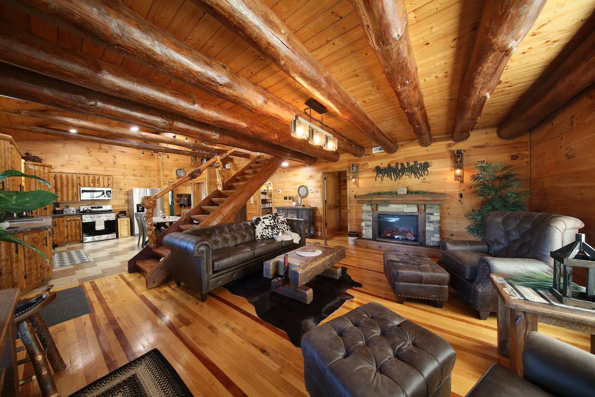 Rustic living room in a log cabin with a stone fireplace, dark leather sofas, and a wooden staircase under large log beams.