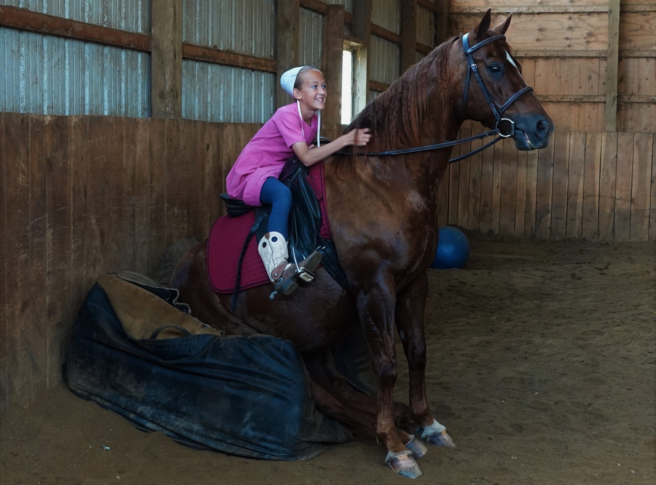 Young girl in a pink shirt rides a brown horse inside a wooden barn/stable.