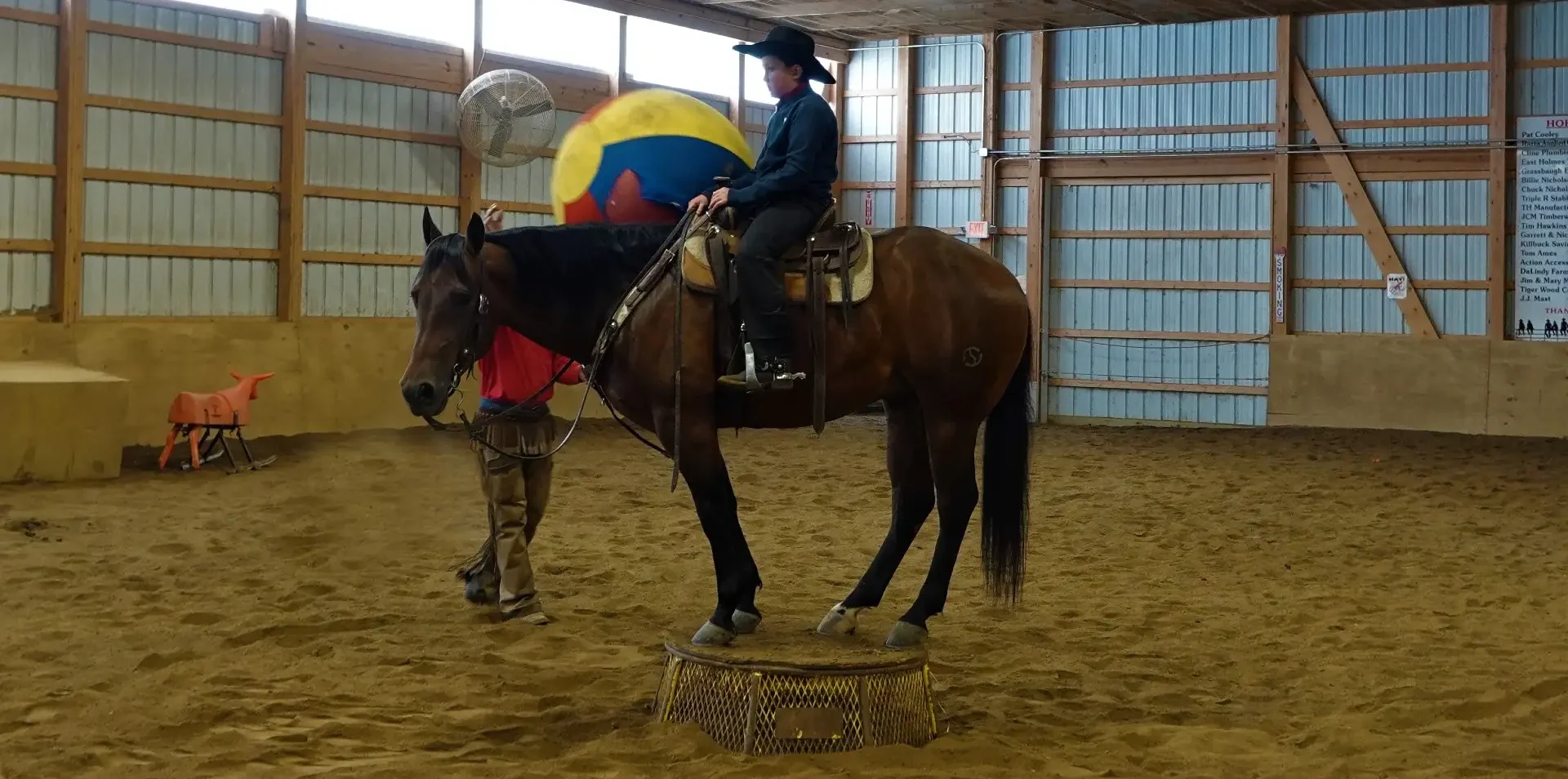 Indoor arena with wooden walls; an American flag hangs from the ceiling as a rider on a brown horse balances a large multi‑colored ball while a trainer stands nearby on a muddy riding surface.