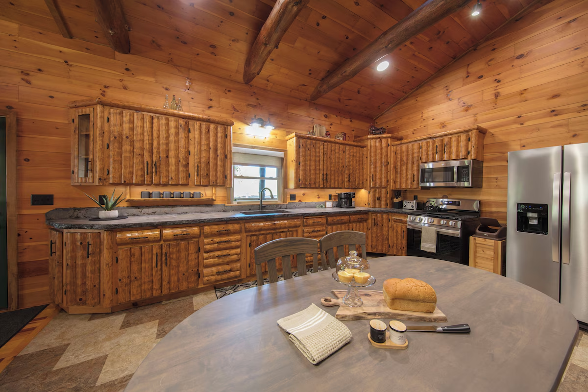 Rustic log cabin kitchen with wooden cabinets, granite counters, and stainless steel appliances around a round dining table with bread and a cake stand in the foreground.