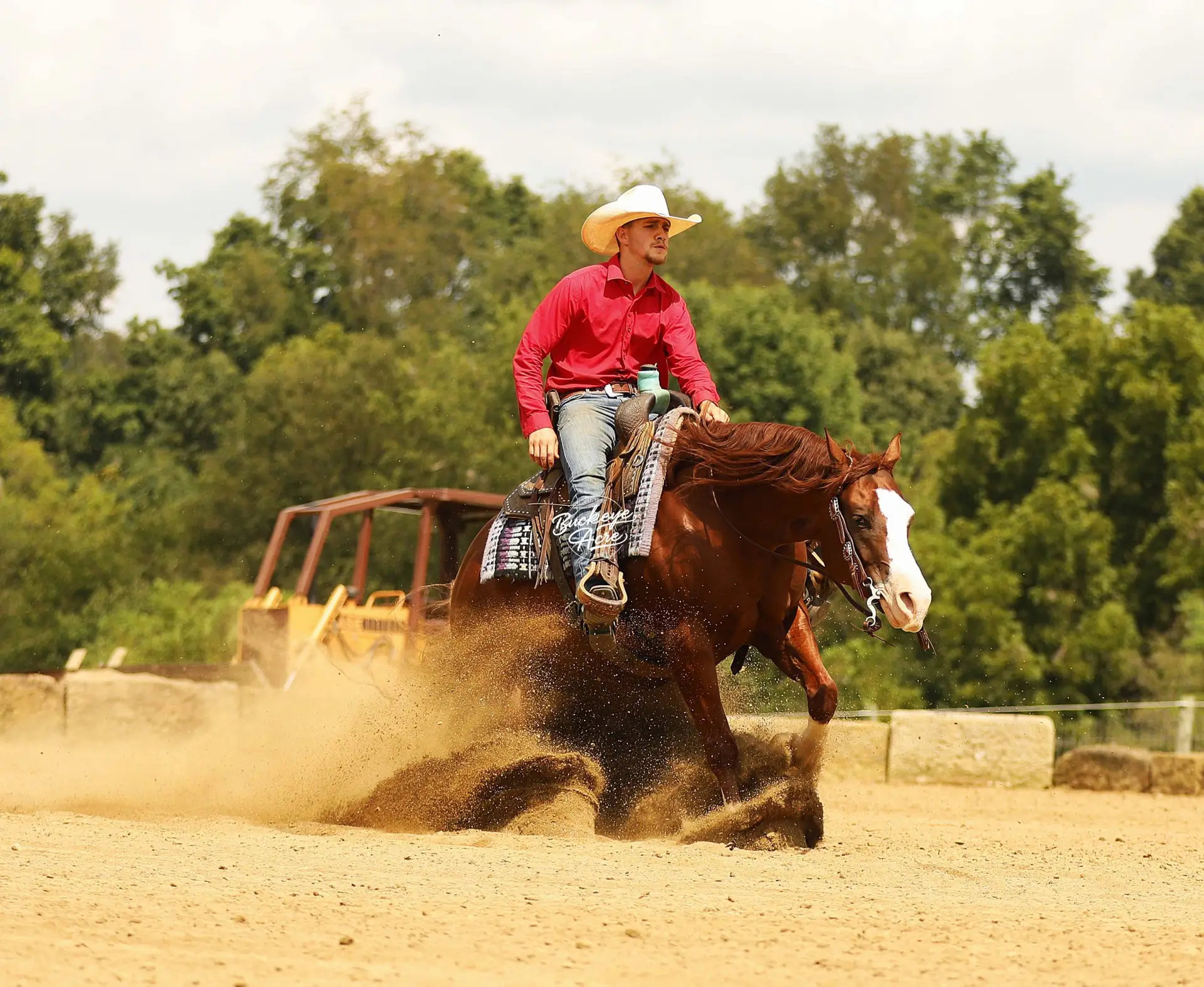 Cowboy in a red shirt and white hat riding a bucking horse in a dusty arena.